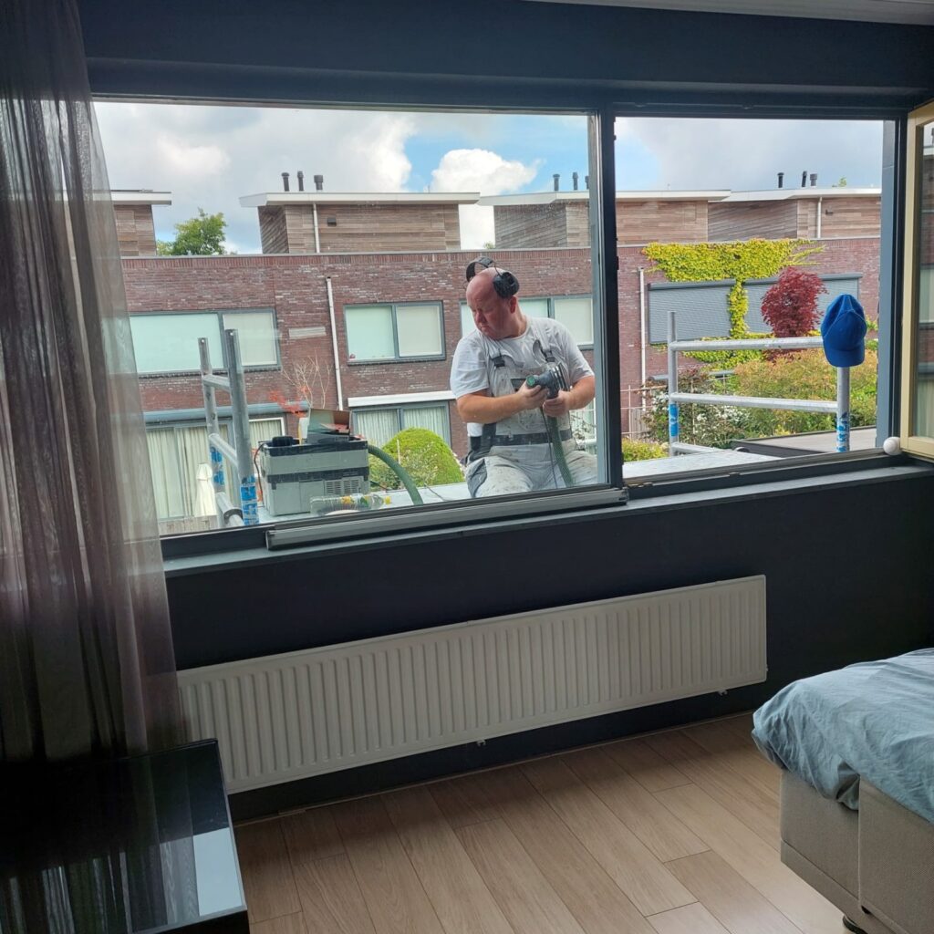 Vakschilder bezig met buitenschilderwerk, gefocust aan het schuren van kozijnen op een steiger.